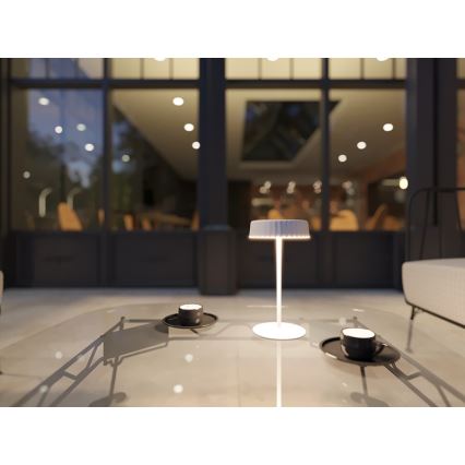 Biela stolná lampa s vrúbkovaným tienidlom svieti na sklenenom stolíku. Na stolíku sú dva tmavé šálky na podšálkach. Pozadie tvorí rozmazané vnútro domu s veľkými oknami.