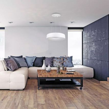 Biela kruhová lampa visí v modernom interiéri. Lampa osvetľuje bielu rohovú sedačku a drevený konferenčný stolík. Okolím je svetlá miestnosť s drevenou podlahou a tmavosivou stenou.