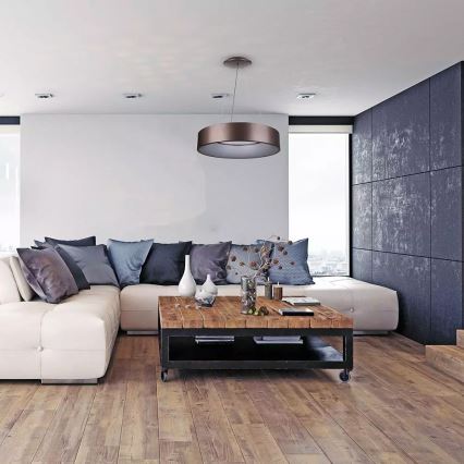 Moderné kruhové svietidlo visí v obývacej izbe. Svietidlo osvetľuje bielu rohovú sedačku s vankúšmi a drevený konferenčný stolík. Izba má drevenú podlahu a tmavosivú stenu.