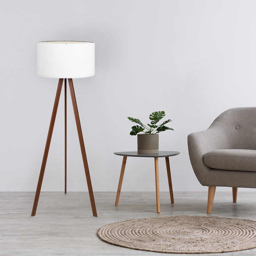 Drevená stojacia lampa s bielym tienidlom stojí v priestore s dreveným stolíkom, na ktorom je kvetináč s rastlinou. Vedľa je sivá sedačka.