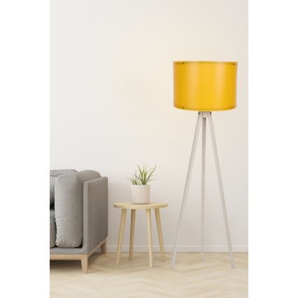 Stojacia lampa s trojnohou podstavou a žltým tienidlom stojí v obývacej izbe vedľa pohovky a nízkeho dreveného stolíka s kvetináčom.