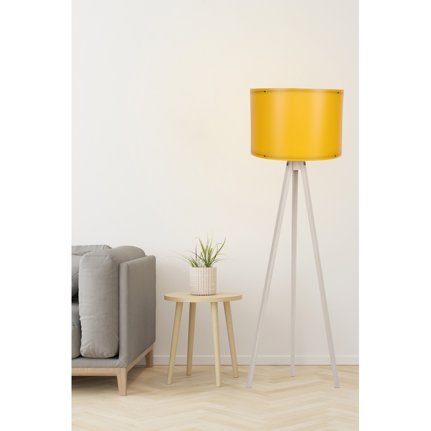 Stojacia lampa s trojnohou podstavou a žltým tienidlom stojí v obývacej izbe vedľa pohovky a nízkeho dreveného stolíka s kvetináčom.