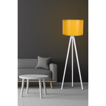 Stojacia lampa s bielym trojnohým stojanom a žltým tienidlom stojí v obývacej izbe pred sivou sedačkou a nízkym bielym stolíkom.