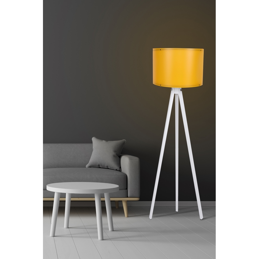 Stojacia lampa s bielym trojnohým stojanom a žltým tienidlom stojí v obývacej izbe pred sivou sedačkou a nízkym bielym stolíkom.