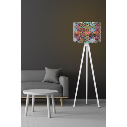 Stojacia lampa s bielym trojnohým podstavcom a valcovým tienidlom s viacfarebným vzorom zloženým z geometrických tvarov a mandál. Lampa stojí v obývacej izbe pred sivou pohovkou a bielym kávovým stolíkom.