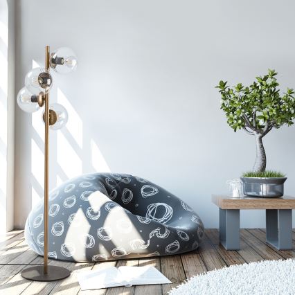 Stojacia lampa s viacerými sklenenými tienidlami stojí pri bezobalovom sedaciemu vaku s abstraktným vzorom. Vedľa je bonsai stromček na drevenom stolíku.