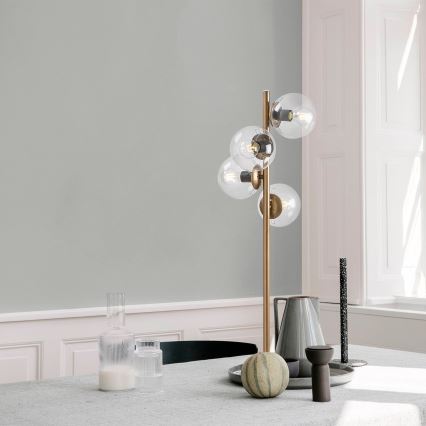 Stolová lampa s viacerými sklenenými guľovými tienidlami stojí na stole. Lampa je zlatá, tienidlá priehľadné. Na stole sú aj džbán, poháre a melón.