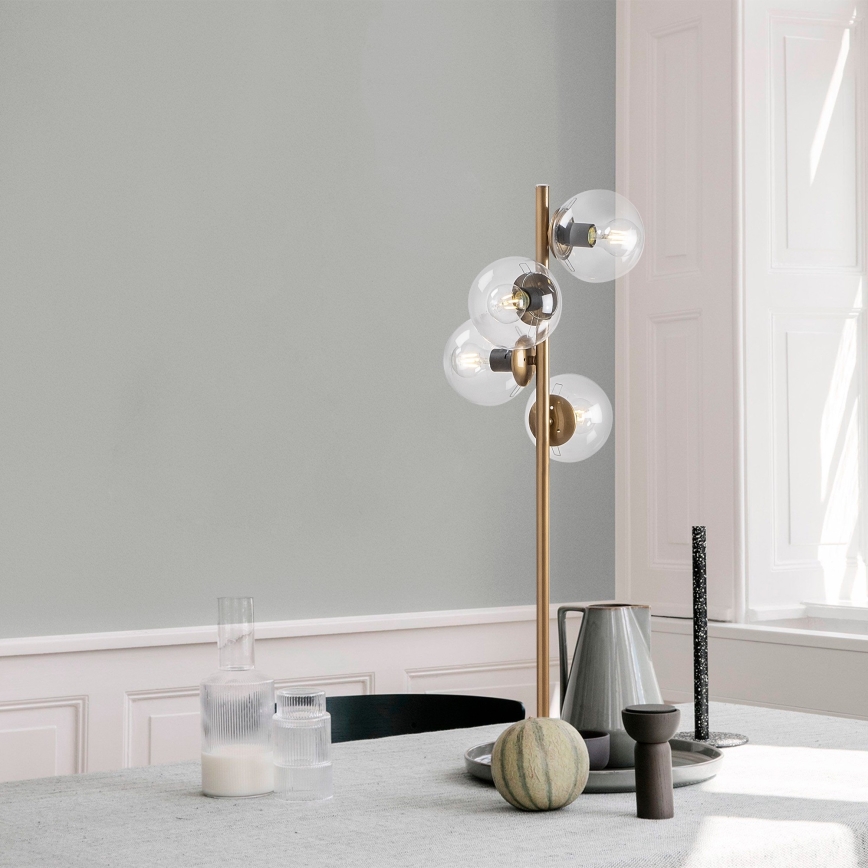 Stolová lampa s viacerými sklenenými guľovými tienidlami stojí na stole. Lampa je zlatá, tienidlá priehľadné. Na stole sú aj džbán, poháre a melón.