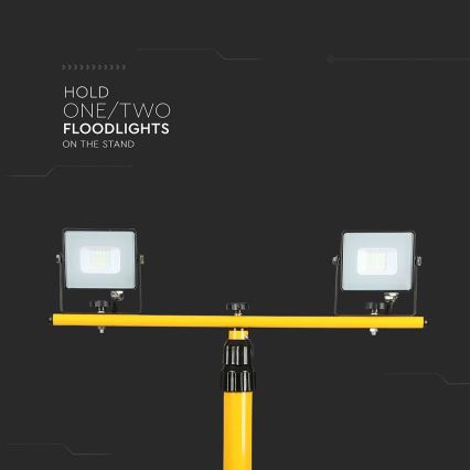 Dve prenosné reflektorové svetlá sú pripevnené na žltej kovovej tyči, ktorá je zasadená do žltého stojana. Text: HOLD ONE/TWO FLOODLIGHTS ON THE STAND.