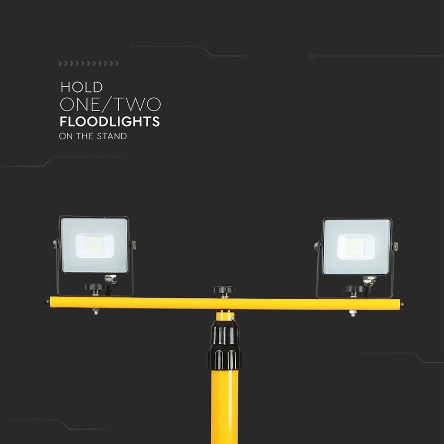 Dve prenosné reflektorové svetlá sú pripevnené na žltej kovovej tyči, ktorá je zasadená do žltého stojana. Text: HOLD ONE/TWO FLOODLIGHTS ON THE STAND.