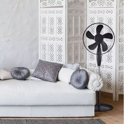 Stolový ventilátor stojí na podlahe pred bielou pohovkou. Ventilátor je čierny s piatimi lopatkami. Pohovka je ozdobená sivými vankúšmi a nachádza sa pred bielou, vyrezávanou priečkou.