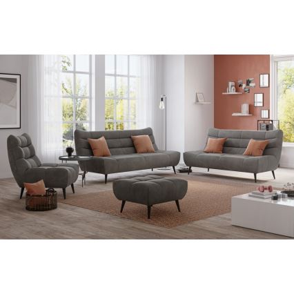 Sivá sedacia súprava, pozostávajúca z trojmiestnej a dvojmiestnej pohovky a kresla, je umiestnená v obývacej izbe na koberci. Súčasťou súpravy je aj taburetka. V pozadí sú okná, stena s poličkou a stolík.