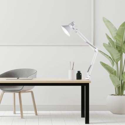 Biela stolová lampa s nastaviteľným ramenom je pripevnená k svetlému drevenému stolu s čiernymi nohami. Na stole sú malé nádoby a vedľa stola stojí sivá stolička a zelená rastlina v kvetináči.