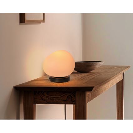 Stolová lampa s guľatým matným skleneným tienidlom svieti na drevenom stole. Lampa je umiestnená v rohu miestnosti. Vedľa nej je drevená miska.
