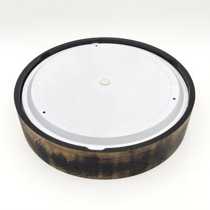 Okrúhly drevený podstavec s bielym diskom v hornej časti, na bielom pozadí. Drevený okraj je tmavo hnedý, čiastočne spálený. Biely disk má v strede malý výstupkový bod a niekoľko malých otvorov.
