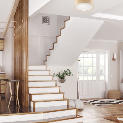 Drevené schody vedú hore v svetlom interiéri domu. Na stene visí dekoratívny štvorcový obraz. V pozadí sa nachádza dvere s oknami.