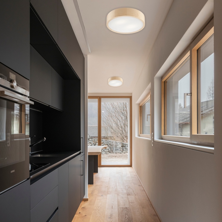 Svietidlo svieti v dlhej chodbe s drevenou podlahou a veľkými oknami. Chodba vedie do kuchyne s tmavou nábytkovou zostavou.