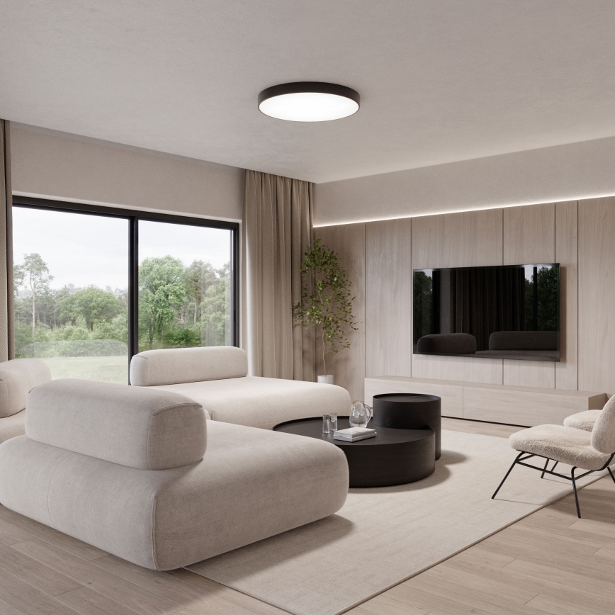 Rohová sedačka stojí v obývacej izbe s výhľadom na zeleň. Sedačka je béžová, pred ňou je nízky čierny konferenčný stolík. V pozadí je drevená stena s televízorom.