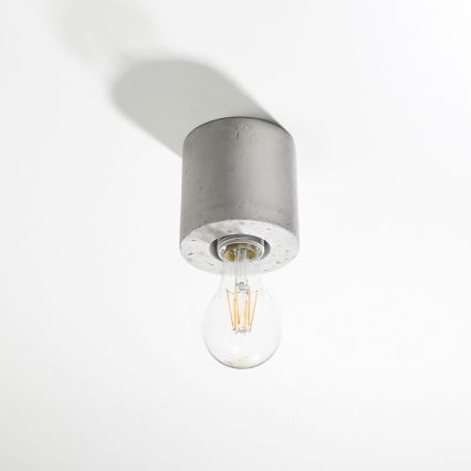 Betónová lampa visí zo stropu. Žiarovka je zasunutá do otvoru v betónovom tiel. Svetlo je zapnuté.