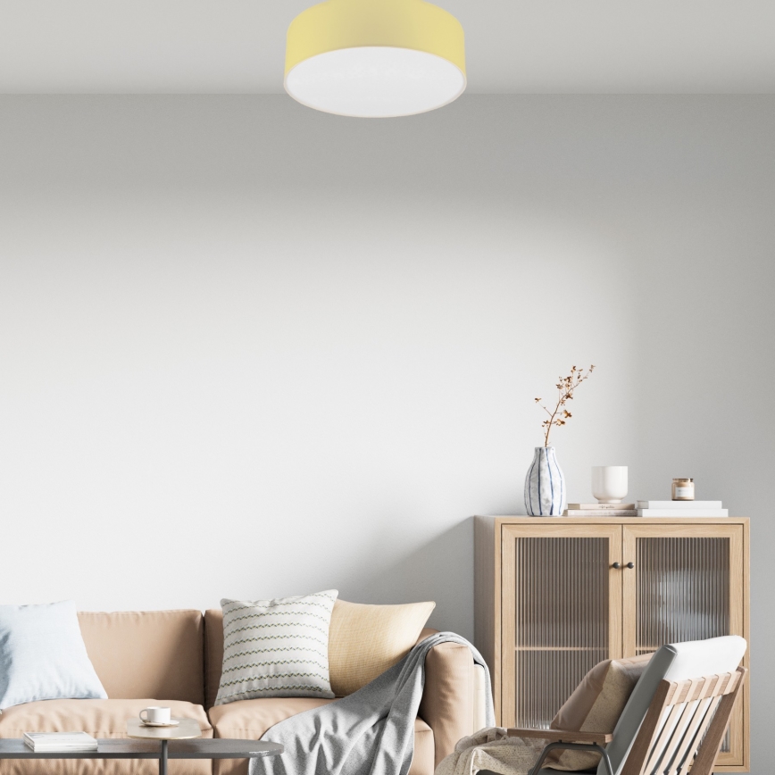 Svetlo s bledžltým tienidlom svieti v obývacej izbe. Svietidlo je pripevnené k stropu nad hnedou sedačkou, drevenou skrinkou a kreslom.