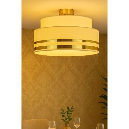 Trojvrstvová stropná lampa s bielym tienidlom a zlatými pásikmi svieti v miestnosti so zlatou tapetou. Lampa je pripevnená k stropu nad jedálenským stolom.