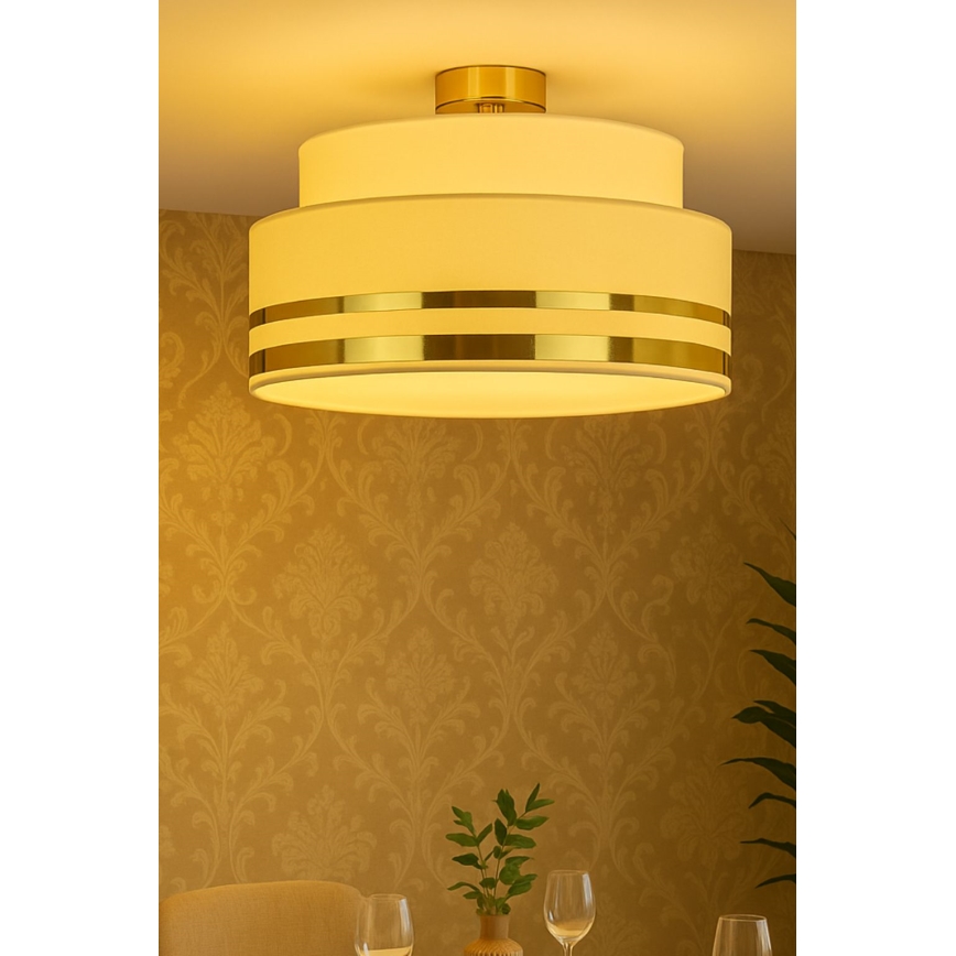 Trojvrstvová stropná lampa s bielym tienidlom a zlatými pásikmi svieti v miestnosti so zlatou tapetou. Lampa je pripevnená k stropu nad jedálenským stolom.