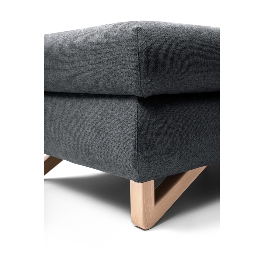 Čalúnená tmavošedá taburetka stojí na svetlých drevených nožičkách tvaru písmena V. Je umiestnená na bielom pozadí.