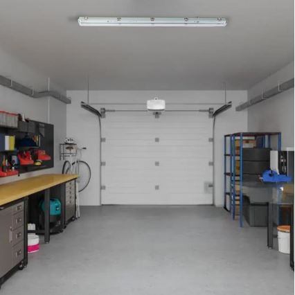 LED svietidlo svieti v garáži. Garáž obsahuje pracovný stôl, bicykel, regály s náradím a pneumatikami, a garážové dvere.