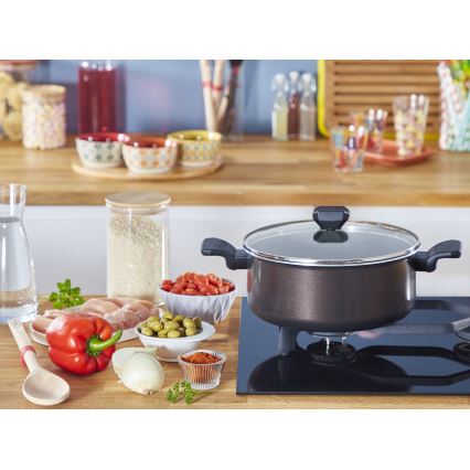 Hrnček s priehľadným vekom sa nachádza na indukčnom variči. Okolo neho sú rozložené suroviny: ryža, kuracie mäso, paprika, cibuľa, olivy a korenie. V pozadí je kuchynská linka s ďalšími potravinami.