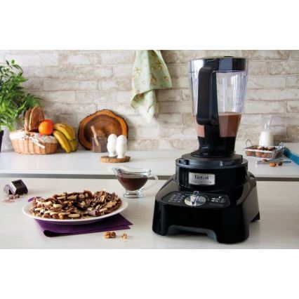 Čierny kuchynský robot Tefal Classic Mix pripravuje nápoj. Nachádza sa na kuchynskej linke pred tanierom s palacinkami, orechmi a omáčkou. Na pozadí je kuchynská stena a ovocie v košíku.