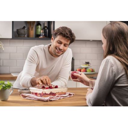 Muž dekorovaním tiramisu malínami. Žena podáva maliny v sklenenej nádobe. Kuchynský stôl s jedlom a riadom.