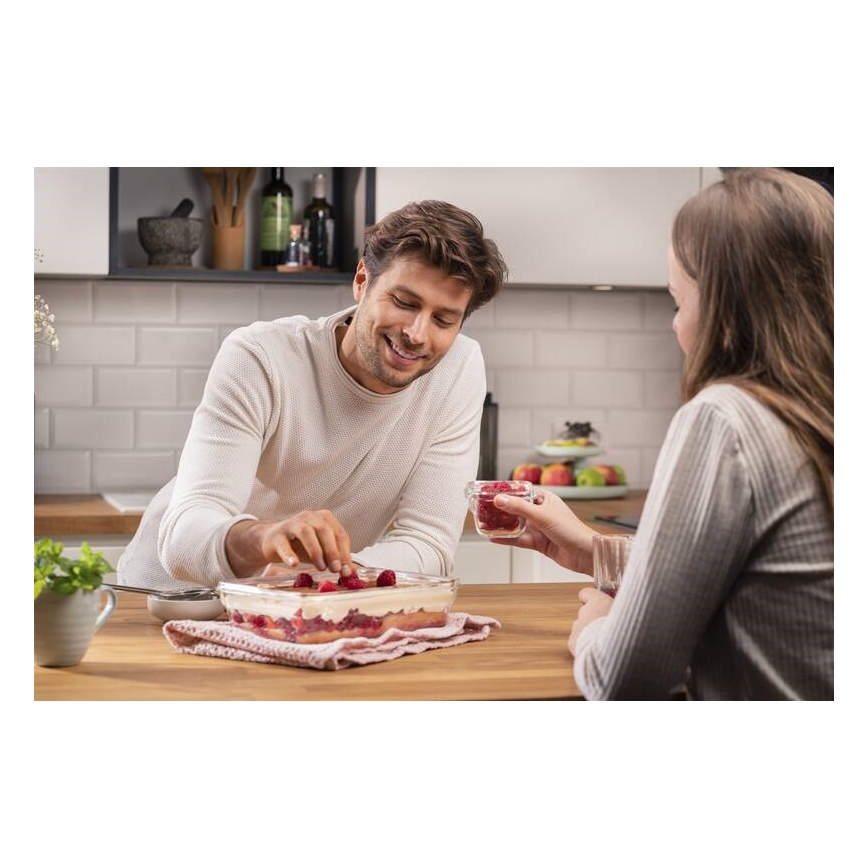 Muž dekorovaním tiramisu malínami. Žena podáva maliny v sklenenej nádobe. Kuchynský stôl s jedlom a riadom.