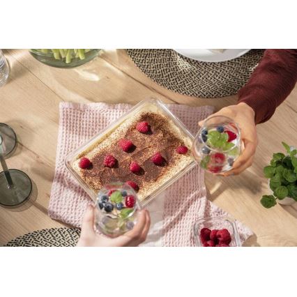 Tiramisu v sklenenej nádobe je stredobodom stola, dve osoby pri stole držia poháre s nápojmi a ovocím. Na stole je tiež ružový obrúsok a miska s malinami.