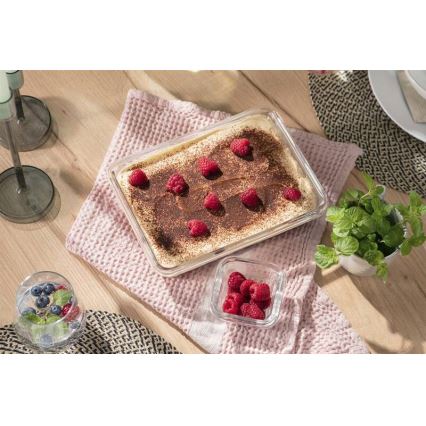 Tiramisu v sklenenej nádobe je umiestnené na svetlom drevenom stole. Na vrchu je posypané kakaom a ozdobené malinami. Vedľa je miska s malinami a mäta.