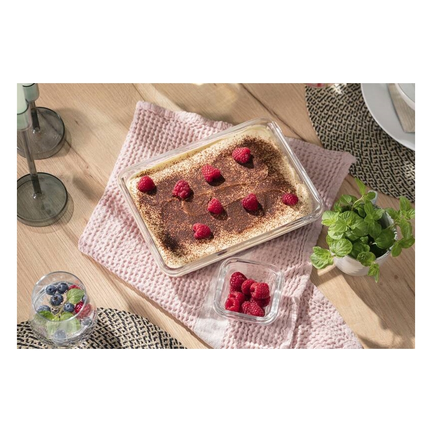 Tiramisu v sklenenej nádobe je umiestnené na svetlom drevenom stole. Na vrchu je posypané kakaom a ozdobené malinami. Vedľa je miska s malinami a mäta.