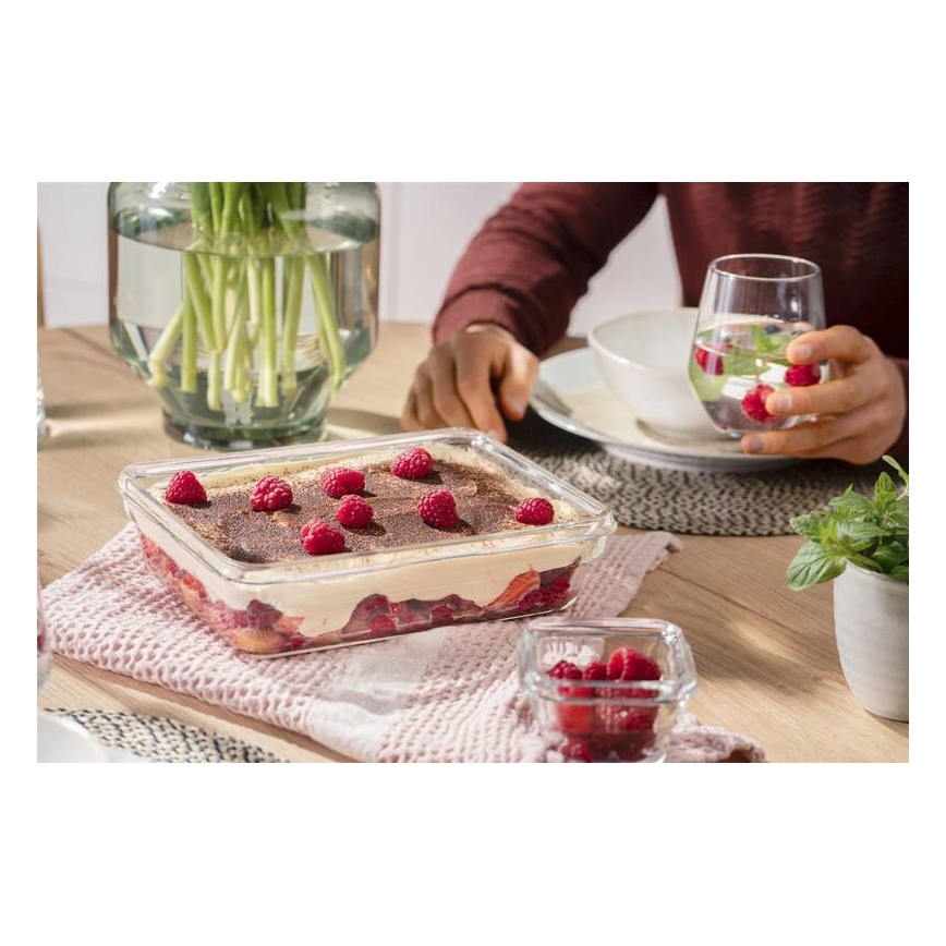 Sklenená nádoba s tiramisu a malinami stojí na stole. Osoba pri stole drží pohár s vodou. Na stole sú aj ďalšie misky s malinami a váza s kvetmi.