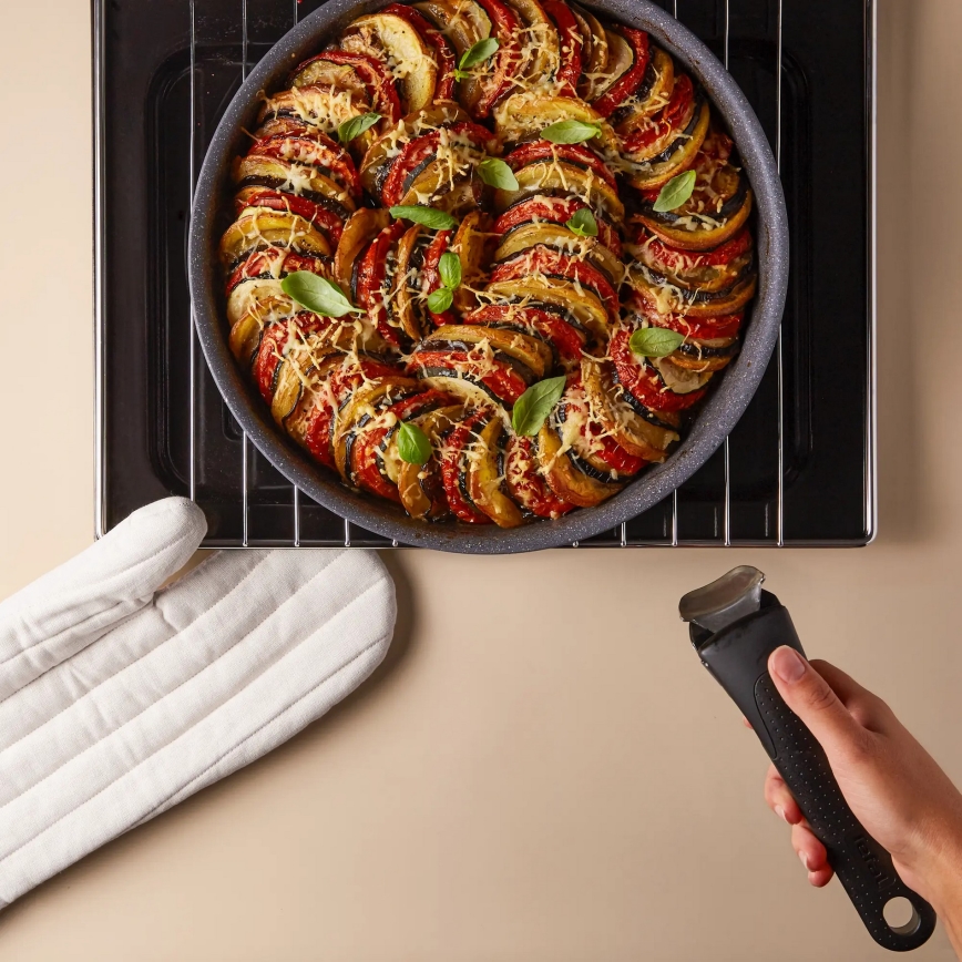 Ratatouille v zapečenej forme sa nachádza na mriežke rúry. Ruka drží kuchynskú rukoväť. Vedľa sa nachádzajú rúrové rukavice na svetlom podklade.