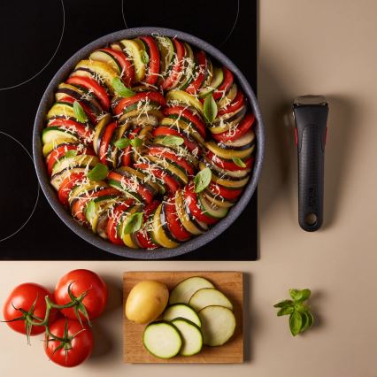 Ratatouille v panvici na indukčnej platni. Je posypané strúhaným syrom a bazalkou. Vedľa sú paradajky, zemiak a nakrájané cukety.