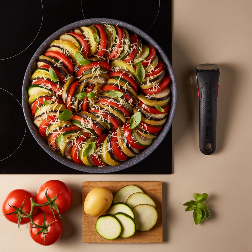 Ratatouille v panvici na indukčnej platni. Je posypané strúhaným syrom a bazalkou. Vedľa sú paradajky, zemiak a nakrájané cukety.