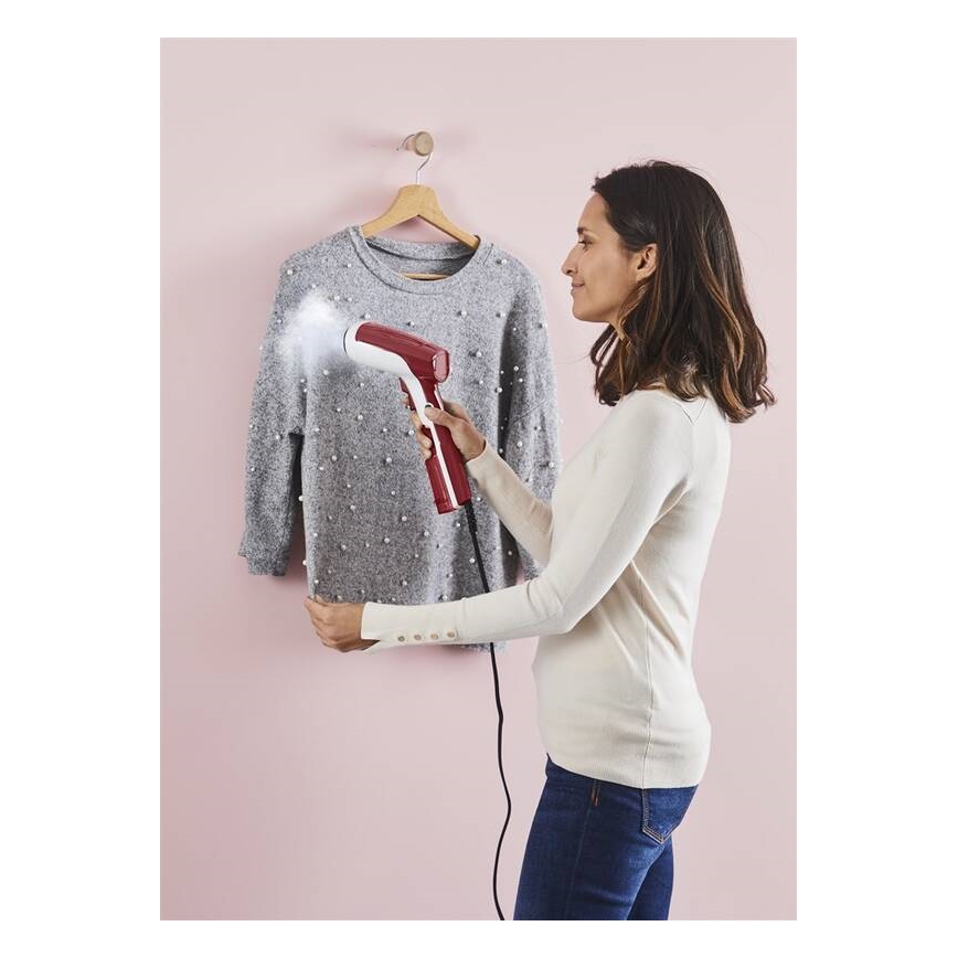 Žena používa ručný parný generátor na vyžehlenie sivého svetra zaveseného na vešiaku. Sveter je zdobený drobnými perličkami. Pozadie je bledoružové.