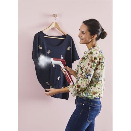 Žena používa ručný parný žehliaci prístroj na vyžehlenie tmavomodrého trička s vyšívanými ananásmi. Trikó je zavesené na vešiaku pred ružovou stenou.