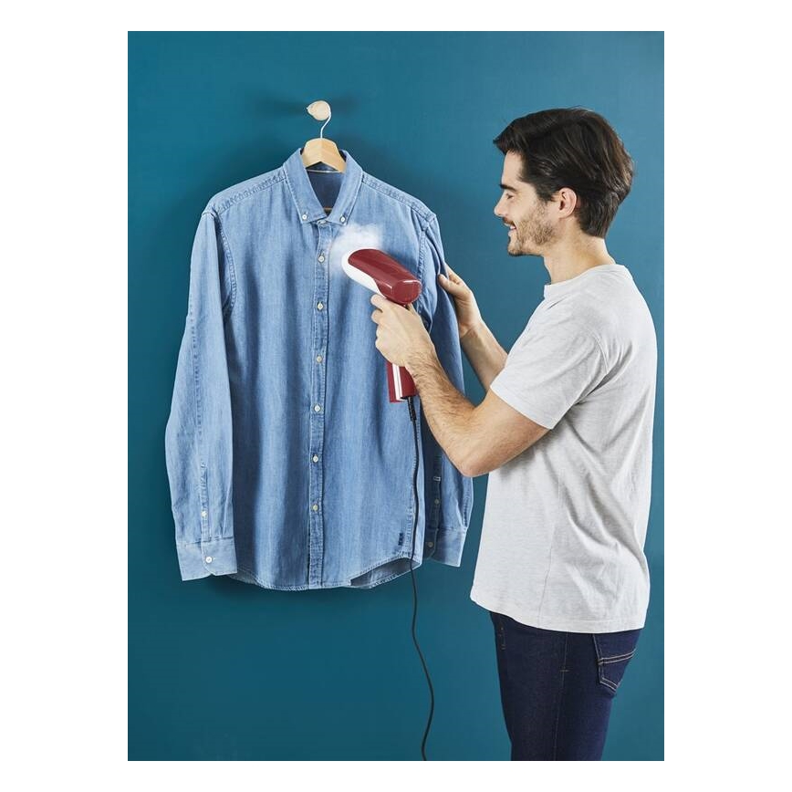 Muž žehlí červeným ručným parným žehličkou džínsovú košeľu zavesenú na vešiaku pred tmavomodrou stenou.