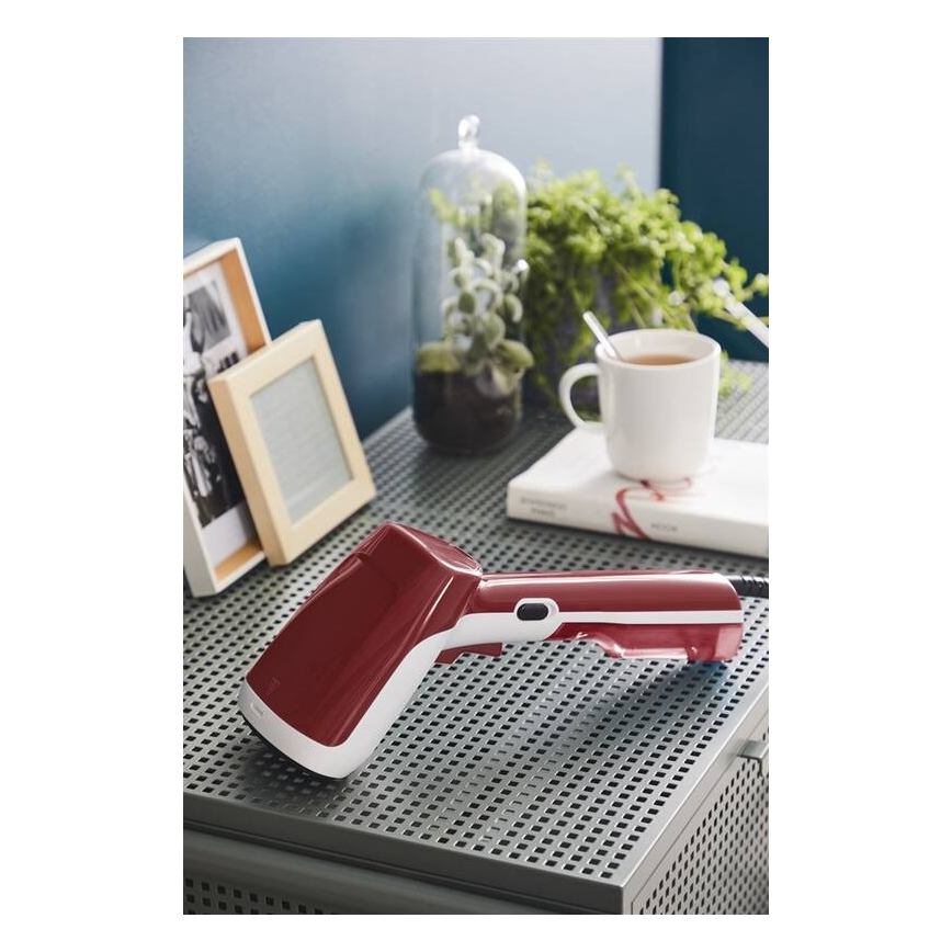 Ručný parný čistič leží na kovovom stolíku. Nachádza sa v červeno-bielej farbe. V okolí sú rámy s fotografiami, kvetináč, šálka a kniha.