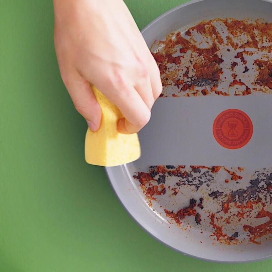 Ruka drží žltú špongiu a čistí špinavú panvicu s hnedými a červenými zvyškami jedla. Na panvici je červený okrúhly nápis: THERMO-ARMA [obrázok presýpacích hodín] THERMO-REGULATING. Panvica leží na zelenej podložke.