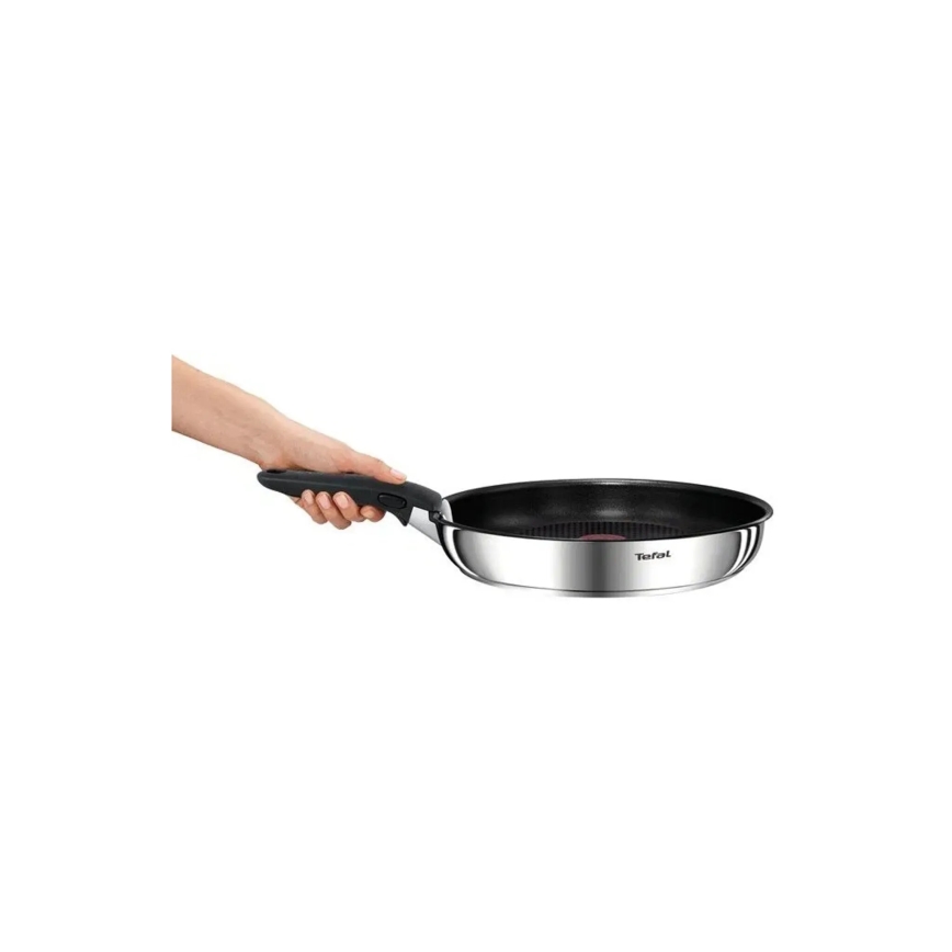 Ruka drží nepriľnavú panvicu Tefal s nerezovou konštrukciou a čiernym dnom. Panvica je držaná za ergonomickú rukoväť. Panvica je umiestnená pred bielym pozadím.