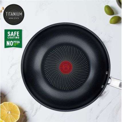 Titánová nepriľnavá panvica s bodkovaným dnom leží na svetlom povrchu. Je označená nápismi „TITANIUM NON-STICK“ a „SAFE COATING NO PFOA NO CADMIUM NO LEAD“. V okolí sú citróny a olivy.