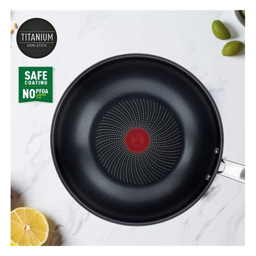 Titánová nepriľnavá panvica s bodkovaným dnom leží na svetlom povrchu. Je označená nápismi „TITANIUM NON-STICK“ a „SAFE COATING NO PFOA NO CADMIUM NO LEAD“. V okolí sú citróny a olivy.