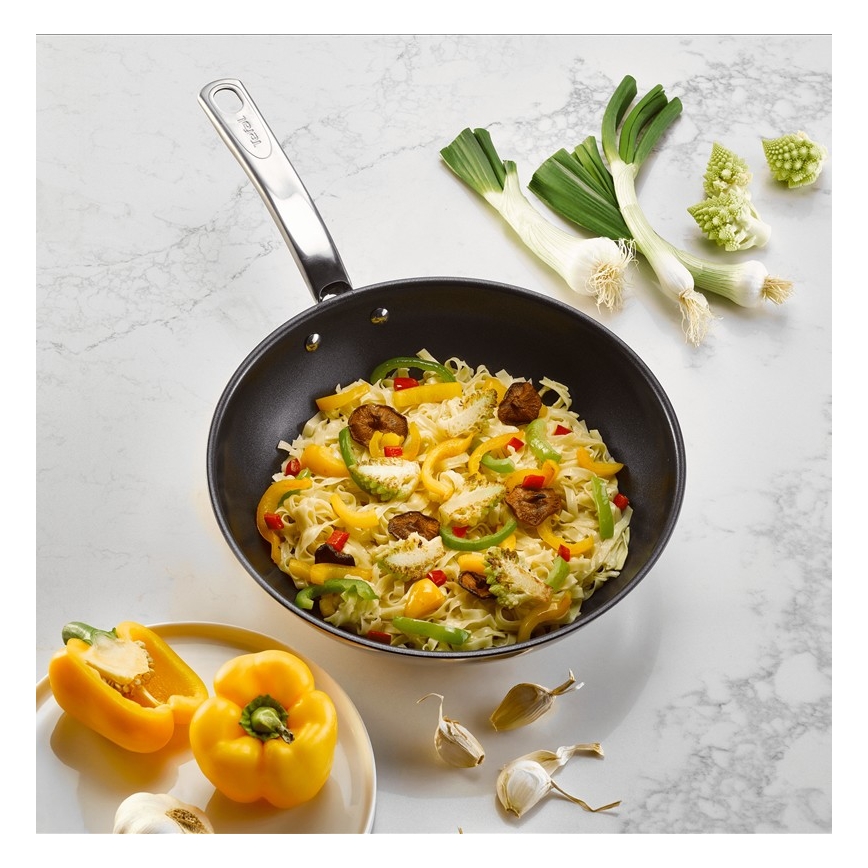 Wok s cestovinovou zmesou a zeleninou, vrátane paprík a romanesco, sa nachádza na mramorovom povrchu spolu s cesnakom a jarnou cibuľkou.