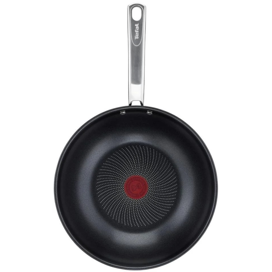 Wok Tefal s kovovou rukoväťou je zobrazený zhora. Jeho vnútorná časť je tmavá s bodkovaným vzorom vychádzajúcim zo stredu, kde je viditeľné červené logo.
