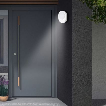 Okrúhle vonkajšie svetlo svieti na sivé dvere. Svetlo je namontované na stene domu priamo vedľa dverí. Pred dverami je kvetináč s levanduľou.